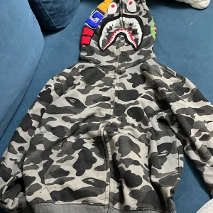 Camouflage hoodie med hajmotiv - Säljer en cool camouflage hoodie från BAPE med ett hajmotiv på huvan. Den har en dragkedja framtill och färgglada detaljer på huvan. Perfekt för streetwear-stilen!