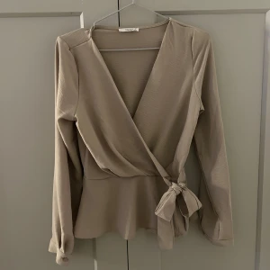 Beige omlottopp från Timming - Säljer en stilren beige omlottopp från Timming. Toppen har långa ärmar och ett knytband i midjan för en smickrande passform. Perfekt för en elegant look.