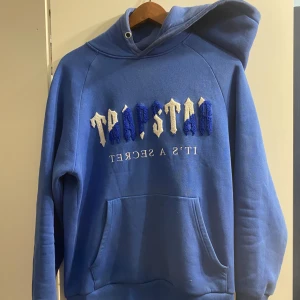 Blå hoodie från Trapstar - Säljer en blå hoodie från Trapstar med texten 'It's a Secret' på framsidan. Hoodien har en klassisk design med känguruficka och en bekväm passform. Perfekt för en avslappnad stil. Storlek S. 