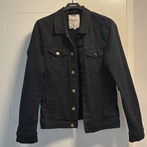 Svart jeansjacka från Jack & Jones - Snygg svart jeansjacka från Jack & Jones med klassisk design. Jackan har knappar framtill och två bröstfickor med lock. Perfekt för en stilren look. 4 fickor