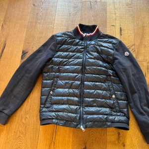 Moncler cardigan - Snygg svart Cardigan från Moncler med långa ärmar och dragkedja. Jackan har ett quiltat mönster och ribbad krage med röd och vit detalj. Perfekt för kyligare sommarkvällar. Hör av er för frågor eller funderingar.