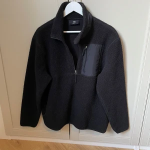 Svart fleecejacka från H&M - Säljer en svart fleecejacka från H&M med halv dragkedja och en praktisk bröstficka med dragkedja. Jackan har en hög krage och är perfekt för kyliga dagar. Den är långärmad och har en mjuk och bekväm känsla.