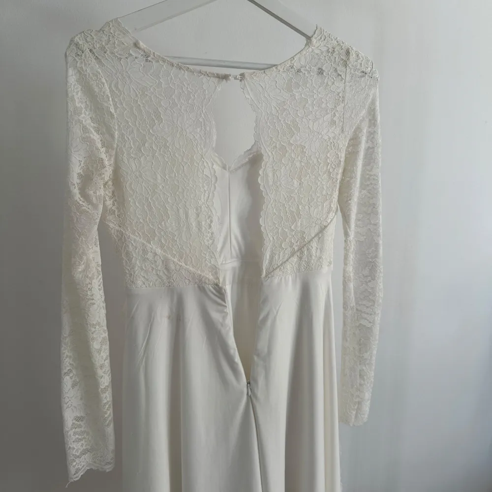Elegant vit klänning med vacker spetsdetalj och lång ärm. Klänningen har en v-ringad design och en mjuk, flödande kjol. Perfekt för speciella tillfällen. Den har även dragkedja på baksidan. . Mekot.