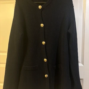 Svart stickad kofta med guldknappar - Säljer en elegant svart stickad kofta med snygga guldknappar framtill. Koftan har långa ärmar och praktiska fickor. Perfekt för att hålla sig varm och stilren under kyligare dagar.