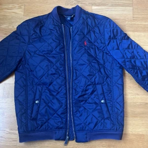 Blå quiltad jacka från Ralph Lauren - Säljer en snygg blå quiltad jacka från Ralph Lauren. Jackan har en dragkedja framtill och två fickor med knappar. Den är långärmad och har en klassisk design med ribbade muddar. Perfekt för kyligare dagar!