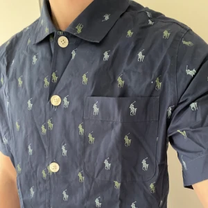Kortärmad Ralph Lauren skjorta - Snygg blå kortärmad skjorta från Ralph Lauren med ett  mönster av små broderade logor i grönt och blått. Skjortan har knappar framtill och en bröstficka. Perfekt för en dag på semestern. Den är helt ny, oanvänd. 