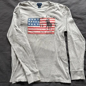 Grå långärmad tröja från Ralph Lauren - Säljer en grå långärmad tröja från Ralph Lauren med ett tryck av en amerikansk flagga och en häst. Perfekt för en avslappnad stil. Tröjan är tillverkad i ett mjukt material som känns skönt mot huden. Storlek 14-16 Passar S