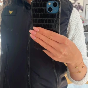 Svart dunväst från Lyle & Scott - Säljer en stilren svart dunväst från Lyle & Scott med dragkedja och det klassiska logotypmärket på bröstet. Perfekt för lager-på-lager under kyligare dagar. Passar både till vardags och mer uppklädda tillfällen.