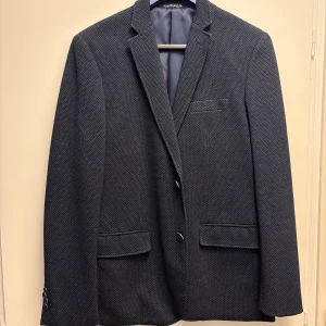 Mörkblå kavaj från Hampton Republic - Snygg mörkblå kavaj från Hampton Republic med diskret mönster. Kavajen har två knappar framtill och fickor med lock. Perfekt för en stilren look.