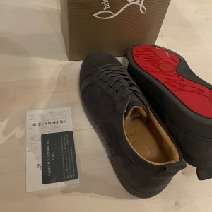 Mörk grå  sneakers från Christian Louboutin - Snygga svarta sneakers från Christian Louboutin med den ikoniska röda sulan. Skorna har snörning och är tillverkade i ett elegant material som ger en lyxig känsla. Perfekta för den som vill ha stil och komfort i ett.