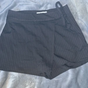 Brun randig shorts kjol från Pull&Bear - Fin brun shorts kjol från Pull&Bear med tunna vita ränder. Kjolen har ett knytband i midjan och en omlottdesign som ger en stilren look. 