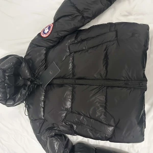 Svart dunjacka från Canada Goose - Säljer en helt ny svart dunjacka från Canada Goose med huva och dragkedja. Jackan har ett quiltat mönster och en logotyp på ärmen. Perfekt för kalla dagar. jackan är i 10/10 skick