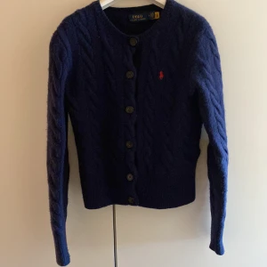Mörkblå kofta från Polo Ralph Lauren - Säljer en snygg mörkblå kofta från Polo Ralph Lauren. Den är stickad med ett klassiskt kabelstickat mönster och har knappar framtill. Koftan är tillverkad i en mjuk ull- och kashmirblandning, vilket gör den både varm och bekväm. Storlek M men passar mer S