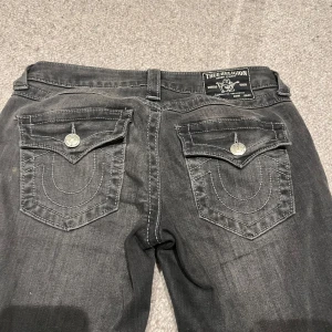 Svarta jeans från True Religion - Snygga svarta jeans från True Religion med bootcut-stil. De har dekorativa bakfickor med knappar och en klassisk femficksdesign. Perfekta för en avslappnad look. Köpta i oktober 2024 och använda ett fåtal gånger, jag säljer pga att jag inte använder dem. Inga defekter. Köpta för 1200kr på Nelly. 