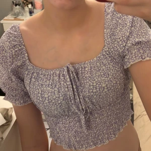 Lila blommig topp - Säljer en söt lila blommig topp med korta puffärmar och knytning framtill. Toppen är croppad och har en smockad nederkant. 