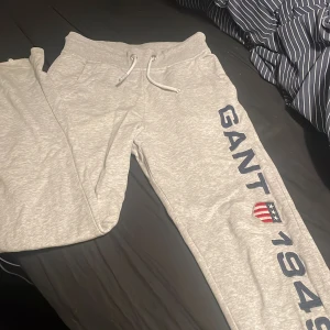 Grå mjukisbyxor från GANT - Säljer ett par grå mjukisbyxor från GANT med tryck 'GANT 1949' och ett broderat emblem på benet. Byxorna har snörning i midjan och är perfekta för en avslappnad stil.