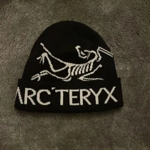 Svart mössa från Arc'teryx - Snygg svart mössa från Arc'teryx med vit logotyp och design. Perfekt för kyliga dagar och ger en stilren look. Passar både till vardags och utomhusaktiviteter.