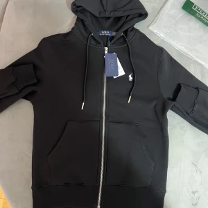 Svart Ralph Lauren Fullzip - Säljer en stilren svart hoodie från Polo Ralph Lauren med dragkedja och broderad logga på bröstet. Tröjan har en klassisk design med känguruficka och justerbar huva. Perfekt för en avslappnad look.