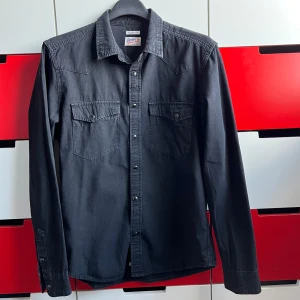 Svart skjorta från Crocker - Snygg svart skjorta från Crocker med klassisk krage och två bröstfickor med knappar. Skjortan har långa ärmar och en normal passform, perfekt för en stilren look. Tillverkad i ett slitstarkt material som ger en bekväm känsla.