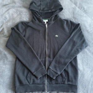 Lacoste Hoodie  - Säljer en stilren svart hoodie från Lacoste med dragkedja och huva. Tröjan har långa ärmar och ribbade muddar vid ärmslut och nederkant. Perfekt för en avslappnad look. Skicket är 9/10. Jag växte ur tröjan innan jag hann använda den så därför säljer jag tröjan. Storlek S - M.                                                         Pris är ej huggit i sten.👍                                            Skriv gärna om du har några frågor!