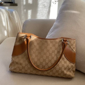 Gucci väska - Säljer en snygg axelväska från Gucci i beige med bruna detaljer. Väskan har det klassiska GG-mönstret. Inuti finns en ficka med dragkedja samt serienummer om någon skulle vara intresserad. Köpt från the vintage bar. Längd: 35,5cm Bredd: 6cm Höjd: 20cm