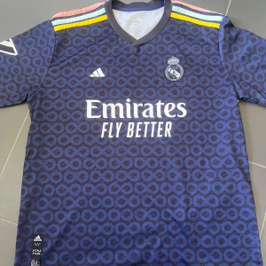 Blå Real Madrid fotbollströja - Snygg blå Real Madrid fotbollströja från Adidas med ett unikt mönster och korta ärmar. Tröjan har klubbens emblem och sponsorlogga på framsidan samt spelarnamnet Benzema och nummer 9 på baksidan. Färgglada detaljer i gult och rosa på axlarna ger en extra touch.