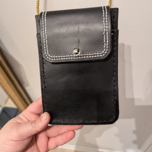 Svart axelväska med guldfärgad rem - Snygg svart axelväska i skinn med kontrasterande vita sömmar och en guldfärgad rem. Väskan har en praktisk knappstängning och en enkel design som passar perfekt för att bära det viktigaste. Perfekt för en stilren look. OBS! Handsydd!