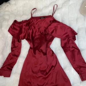 Vinröd klänning från Boohoo - Snygg vinröd klänning från Boohoo med offshoulder-design och volangdetaljer. Klänningen har en elegant och glansig finish, perfekt för en kväll ute. Den är tillverkad i ett mjukt och följsamt material med smala axelband.