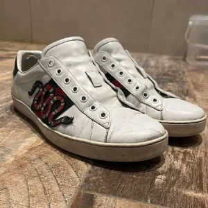 Vita sneakers med ormmotiv från Gucci - Snygga vita sneakers från Gucci med ett coolt ormmotiv i rött och svart på sidan. Skorna har en rund tå och detaljer i rött och grönt på hälen. Perfekta för att ge din outfit en lyxig touch.