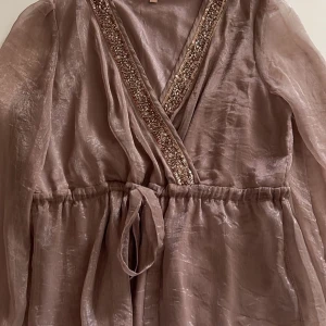 Champagnefärgad blus med paljettdetaljer - Elegant champagnefärgad blus med v-ringning och paljettdetaljer längs kanten. Blusen har långa ärmar och ett knytband i midjan för en smickrande passform. Perfekt för en stilfull look.