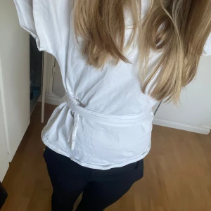 Vit t-shirt med knytband - Säljer en stilren vit t-shirt med korta ärmar och ett knytband i midjan för en snygg passform. Den här även jättesnygga armar som sticker ut lite!