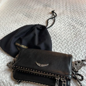 Svart axelväska från Zadig & Voltaire - Snygg svart axelväska i skinn från Zadig & Voltaire med silverdetaljer och en kedjerem. Väskan har en dragkedja på baksidan och ett stilrent emblem framtill. Perfekt för att ge din outfit en edgy touch.