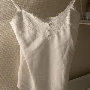 Vit linne med spetsdetaljer - Säljer ett stilrent vitt linne med vackra spetsdetaljer vid halsringningen. Linnet har smala axelband och en knappdetalj framtill. Perfekt för varma dagar eller som ett lager under en kavaj. Är ej använd.