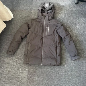 Svart pufferjacka från Sail Racing - Snygg svart pufferjacka från Sail Racing med huva och dragkedja framtill. Jackan har en stilren design med en bröstficka och är perfekt för kyligare väder.