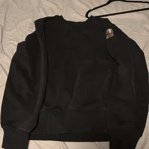Svart fleece tröja från Parajumpers - Säljer en svart fleece tröja från Parajumpers med en liten broderad detalj på ärmen. Tröjan har ribbade muddar vid ärmslut och nederkant, vilket ger en skön passform. Perfekt för kyliga dagar!