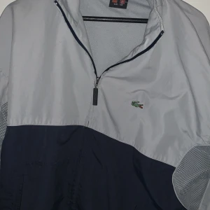 Grå och marinblå vindjacka från Lacoste - Säljer en riktigt skön vindjacka från Lacoste, en äldre modell. Rätt väl använd men fortfarande super bra skick