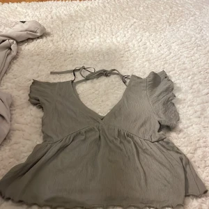 Grå blus med volangdetaljer - Säljer en söt grå blus med volangdetaljer och knytning i ryggen. Blusen har korta ärmar och en v-ringad design. Perfekt för en stilren look.