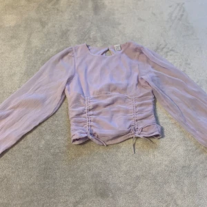 Ljuslila blus med långa ärmar - Säljer en söt ljuslila blus med långa ärmar och smockdetaljer på framsidan. Blusen har en knytning i midjan och en liten öppning bak i nacken. Perfekt för en stilren look!