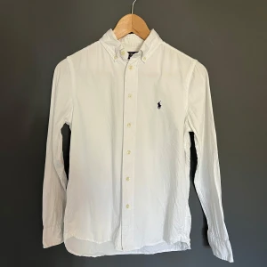 Vit skjorta från Ralph Lauren - Säljer en klassisk vit skjorta från Ralph Lauren med lång ärm och knappstängning för den har blivit för liten. Skjortan har en broderad logotyp på bröstet och är i slim fit. Det är storlek 14 men sitter som en xs. Den är i 9/10 skick. Perfekt för en stilren look.