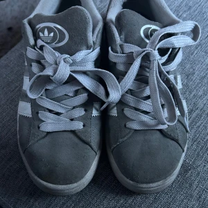 Grå sneakers från Adidas campus 00 - Säljer ett par grå sneakers från Adidas Campus 00 med klassiska vita ränder och snörning. Skorna har en rund tå och är tillverkade i mocka. Perfekta för en avslappnad stil. Storlek 37 1/3. Skorna är använda och har tecken på användning men de är väl omhändertagna. Hör av dig vid intresse💕