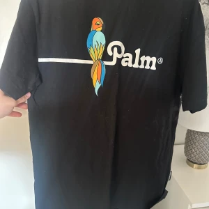 Svart t-shirt från Palm Angels - Snygg svart t-shirt från Palm Angels med en färgglad papegoja och texten 'Palm' på framsidan. Tillverkad i 100% bomull för en skön och bekväm känsla. Perfekt för en avslappnad stil.