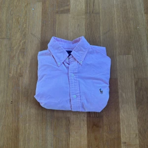 Rosa skjorta från Ralph Lauren - Säljer en stilren rosa skjorta från Ralph Lauren med klassisk krage och knappar framtill. Skjortan har en broderad logotyp på bröstet och är perfekt för en snygg och avslappnad look.