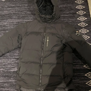 Svart dunjacka från Sail Racing - Säljer en svart dunjacka från Sail Racing med huva och dragkedja. Jackan har en stilren design med logotyp på bröstet och en ficka på ärmen. Perfekt för kalla dagar. Byte går också, sänker pris vid snabb affär