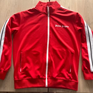 Palm angels  - Snygg röd träningsjacka från Palm Angels med vita detaljer och dragkedja. Jackan har långa ärmar med vita ränder längs sidorna och en broderad logotyp på bröstet. Perfekt för en sportig look!