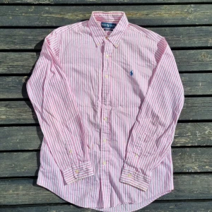 Rosa randig skjorta ralph lauren stl M - Säljer en stilren rosa och vit randig skjorta från Ralph Lauren. Skjortan har långa ärmar och en klassisk krage med knappar. Den är broderad med den ikoniska logotypen i blått på bröstet. Perfekt för en snygg och avslappnad look.