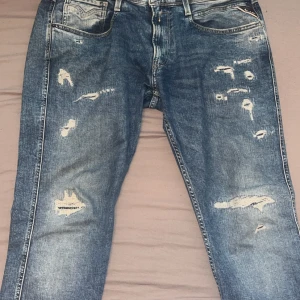 Blå jeans från Replay - Snygga blå jeans från Replay med slitna detaljer och en slim fit passform. Perfekta för en avslappnad stil. Modellen är Anbass och de har en klassisk femficksdesign. Som nya
