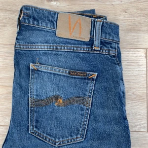 Blå jeans från Nudie Jeans - Snygga blå jeans från Nudie Jeans med klassisk femficksdesign och orange sömmar. De har en slim passform och är perfekta för en avslappnad stil. Märkeslogga på bakfickan ger en extra touch. W29 L30 ett litet hål men inget som märks.