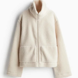 Beige teddyjacka med dragkedja H&M - Mysig beige teddyjacka med dragkedja och hög krage. Perfekt för kyliga dagar med sina långa ärmar och två praktiska fickor framtill. Jackan har en mjuk och fluffig textur som ger en avslappnad look.