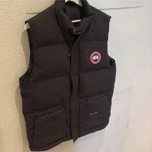 Svart dunväst från Canada Goose - Säljer en svart dunväst från Canada Goose med deras ikoniska märke på bröstet. Västen har praktiska fickor och stängs med dragkedja och knappar. Perfekt för kyliga dagar.