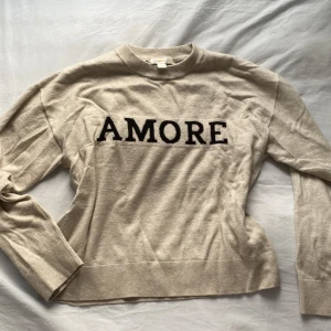 Beige tröja med text från Gina Tricot - Säljer en beige tröja från Gina Tricot med svart text 'AMORE' på framsidan. Tröjan har långa ärmar och en rund halsringning. Perfekt för en avslappnad stil.